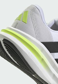 Zapatilla deportiva gris claro con parte superior de malla, acentos en negro, detalle en el talón de color amarillo brillante y suela texturizada etiquetada "CLOUDFOAM" para amortiguación.