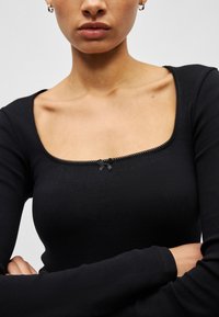 Moteris dėvi juodą ilgomis rankovėmis viršutinę dalį su kvadrato formos iškirpte ir maža dekoratyvine kaspinėliu centre. Rankos sukryžiuotos.