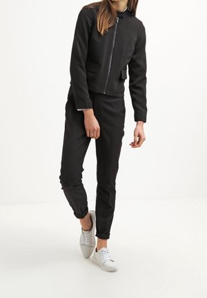 Donna che indossa una giacca nera con zip e tasche a filetto, pantaloni neri con risvolti e sneakers bianche, in piedi davanti a uno sfondo bianco neutro.