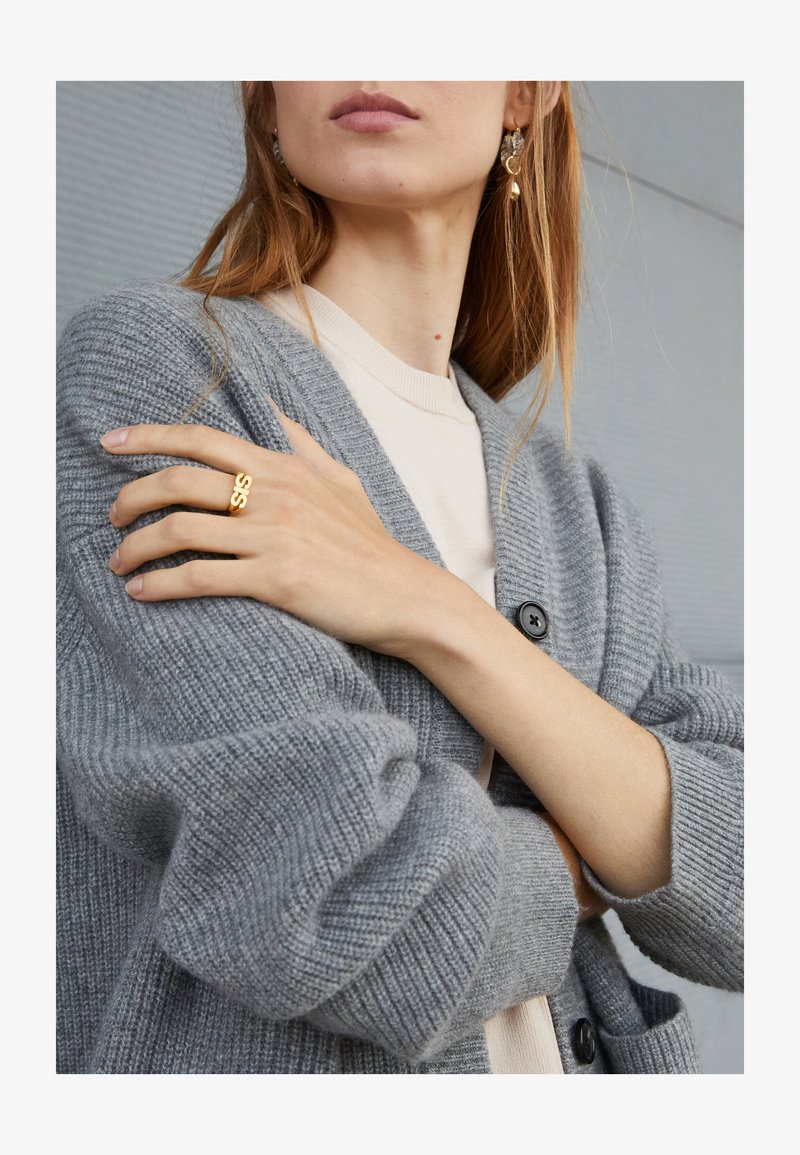 Cardigã cinza tricotado com textura canelada e mangas oversized, combinado com uma blusa creme. Anel dourado com o texto "SIS" e brincos decorativos.