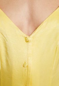 Robe en satin jaune avec un décolleté en V, des coutures verticales et deux boutons assortis sur le devant. Texture douce avec un léger éclat.