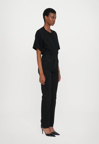 Black cotton t-shirt with short sleeves, paired with high-waisted black jeans and pointed black heels, shown from the side.