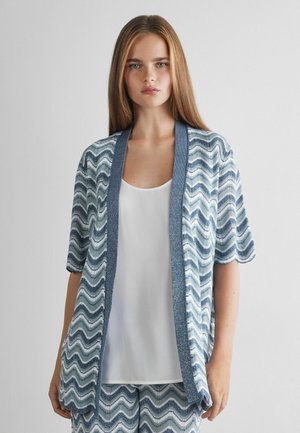 Junge Frau trägt eine kurzärmelige, blau-weiße, wellenförmige Strickjacke über einem weißen Top und steht vor einer schlichten hellen Wand.