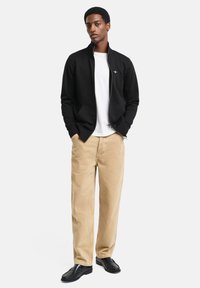 Schwarze Zip-Jacke, weißes Hemd, bequeme beige Weite-Hosen und schwarze Slipper. Glattes Material, lockere Passform und minimalistisches Design.