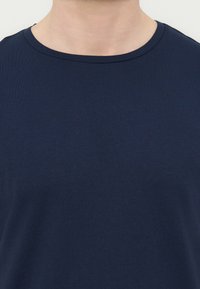 Κοντινό πλάνο ενός ατόμου που φοράει μονόχρωμο μπλε σκούρο τηλεκοντρόλ με στρογγυλή λαιμόκοψη, που δείχνει τον λαιμό και την άνω περιοχή του στήθους.