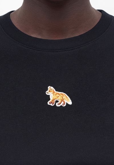 Κοντινό πλάνο ενός ατόμου που φοράει μαύρο πουκάμισο με μικρή κεντημένη πορτοκαλί και λευκή αλεπού στο στήθος.