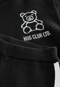 Sort strikket stof med hvidt broderet teddy-bear og teksten "HUG CLUB LTD.", der viser ribbet talje og hvid stribet detalje.