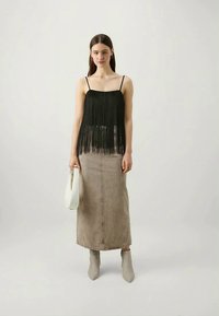 Top nero con frange e spalline sottili, abbinato a una gonna maxi in denim beige. La modella indossa stivali alla caviglia grigio chiaro e tiene una borsa bianca.