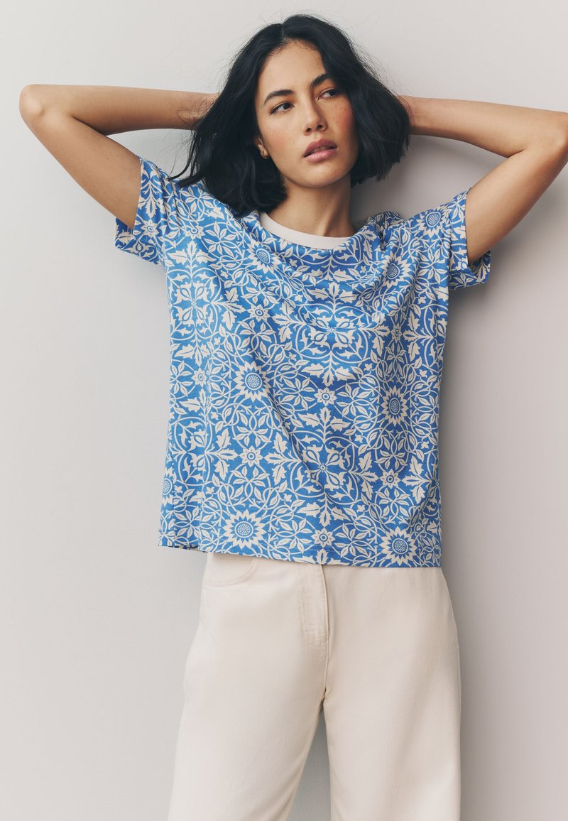 Blaues Kurzarmshirt mit weißem Blumenmuster. Der Stoff wirkt leicht und atmungsaktiv, mit rundem Ausschnitt und lockerem Schnitt.