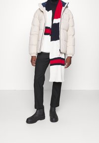 Hombre vestido con chaqueta de plumas beige, suéter de cuello alto negro, pantalones negros, botas negras, y una larga bufanda de punto blanca, roja y negra, de pie contra una pared blanca.