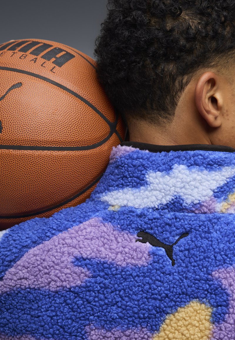 Maglione in pile blu, viola e crema, con motivo a nuvole e logo Puma nero, posizionato accanto a un pallone da basket.