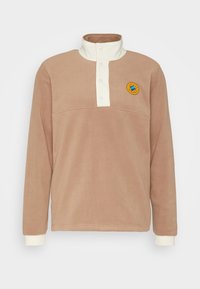 Fleecepullover i ljusbrun med en vit morotskrage och knappslå. Har en broderad cirkulär patch på bröstet.