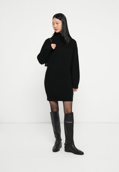 Jeune femme portant une robe pull côtelée noire, des collants noirs, des bottes en cuir noir jusqu'aux genoux, et portant un sac à bandoulière noir.