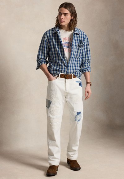 Νέος άνδρας που φοράει ένα μπλε καρό πουκάμισο πάνω από ένα γραφικό μπλουζάκι, λευκό παντελόνι με μπαλώματα, καφέ ζώνη, καφέ μπότες και ένα ρολόι χειρός, στέκεται με τα χέρια στις τσέπες.