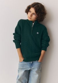 Tamsiai žalti megzti megztinis su pynimo raštais, užtrauktuko apykakle ir mažu logotipu krūtinėje; derinamas su šviesiai mėlynomis džinsais.