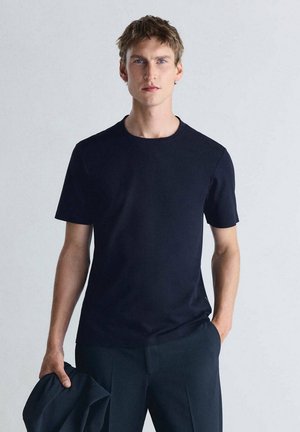 Jeune homme aux cheveux châtain clair portant un t-shirt noir uni à manches courtes et un pantalon foncé, tenant une veste sombre dans une main.