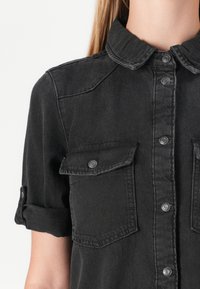 Zwarte denim shirt met korte mouwen, knoopsluiting aan de voorkant, twee borstzakken en opgerolde mouw cuffs. Voorzien van metalen drukknoopsluitingen en een kraagontwerp.