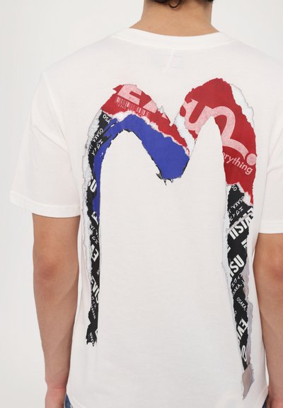 T-shirt blanc avec un grand design graphique au dos, comprenant des blocs de couleurs rouge, bleu et noir ainsi qu'une texture de papier déchiré.