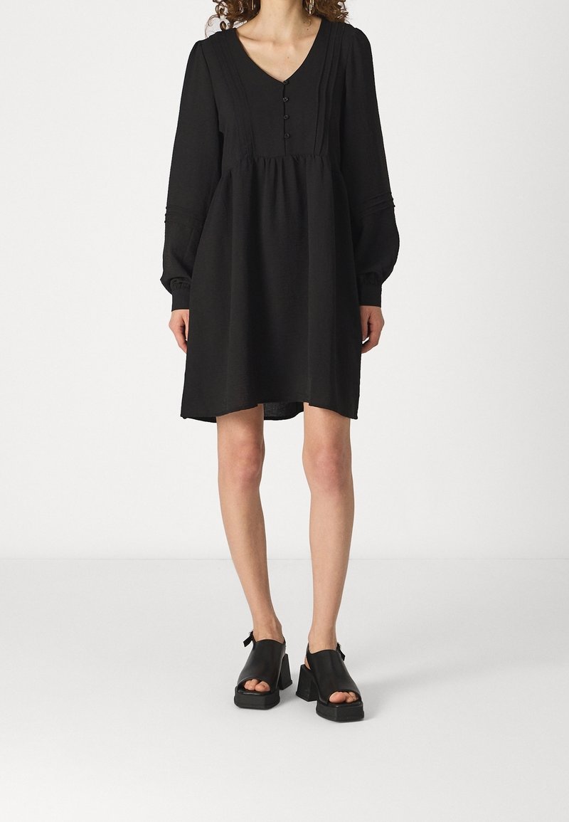 Robe noire à manches longues avec un décolleté en V, détail de boutons et taille froncée. Chaussures plates à talon carré. Tissu simple et texturé.
