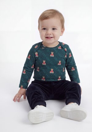 Tout-petit aux cheveux châtain clair assis, portant un t-shirt vert à manches longues avec des ours en peluche, pantalon foncé et chaussures blanches, souriant et regardant devant.
