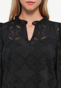 Femme portant un haut noir à manches longues avec des motifs en dentelle florale et un petit col en V à bords froncés.