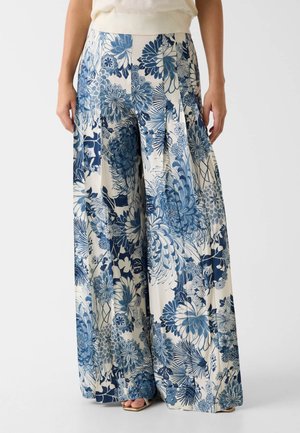 Pantalones de pierna ancha con estampado floral azul y blanco, llevados con top beige sin mangas, manos relajadas a los lados, sandalias beige abiertas en los dedos visibles.