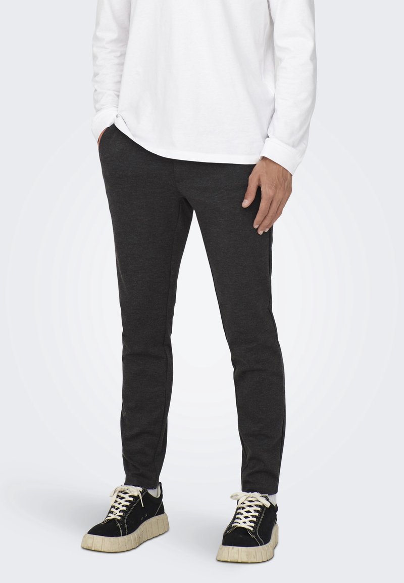 Man som bär mörkgrå slim-fit byxor, vit långärmad skjorta och svarta sneakers med beige sulor, står med händerna i fickorna.