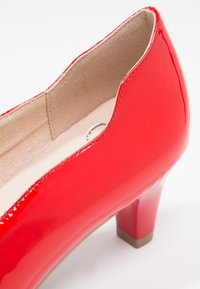 Chaussure à talon haut en cuir verni rouge, avec un bout rond, un talon court et une finition lisse et brillante. L'intérieur est doublé d'un tissu type suède de couleur beige clair.