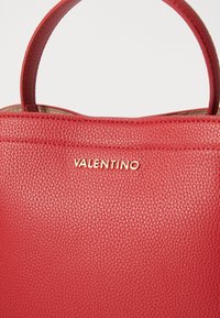 Geantă de piele roșie cu textură granulată, având un design simplu și mânere superioare. Logo-ul auriu "Valentino" afisat proeminent pe față.