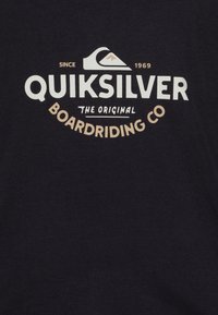 Svart bomull T-skjorte med et fremtredende hvitt og beige Quiksilver-logo med et fjell- og bølgedesign. Teksten inkluderer "Siden 1969" og "Det originale boardriding-selskapet."