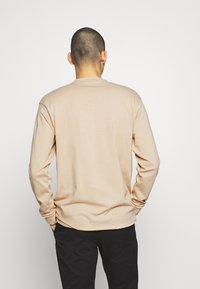 Béžové tričko s dlouhým rukávem, kulatým výstřihem a rovním lemem. Měkká tkanina bez švů na ramenou a s minimem švových detailů.