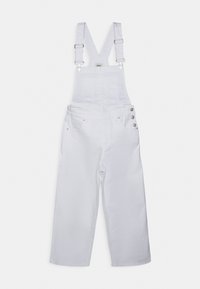 Bílé džínové kšandy s nastavitelnými křížovými popruhy, přední kapsou na hrudi, bočními knoflíky a širokými nohavicemi na jednobarevném bílém pozadí.