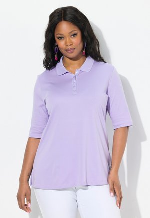 Vrouw in een lichtpaarse poloshirt met korte mouwen en witte broek, met lang donker haar en paars- en roze hangende oorbellen.