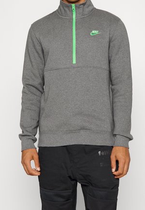 Mann trägt einen grauen Langarm-Nike-Pullover mit einem Viertelreißverschluss in Neon-Grün und Logo, kombiniert mit schwarzen Hosen mit Netz-Einsätzen an den Knien.