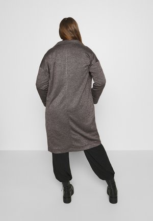 Frau mit langem Haar, die einen strukturierten grauen kniehohen Mantel, schwarze weite Hosen und schwarze Stiefeletten trägt und mit dem Rücken zur Kamera vor einem schlichten Hintergrund steht.