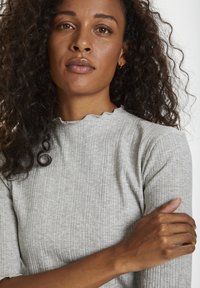 Grå randig topp med scalloped halsringning och korta ärmar, bärs av en modell med lockigt hår. Tyget verkar mjukt och texturerat.