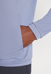 Μπλε αθλητική μπλούζα με μακριά μανίκια και υφή κυψελοειδούς σχεδίασης. Διαθέτει πλαϊνή τσέπη και αντίθετη γκρίζα ραφή κατά μήκος του μανικιού.