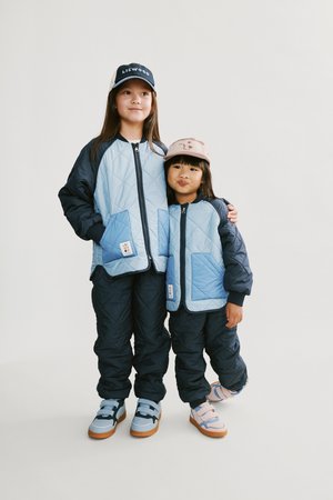 Kaksi lasta pukeutuvat yhteensopiviin vaaleansinisiin ja tummansinisiin tikattuihin takkeihin, joihin kuuluu samansävyiset tummansiniset housut ja värikkäät kengät. Asut viimeistelevät lippalakit.