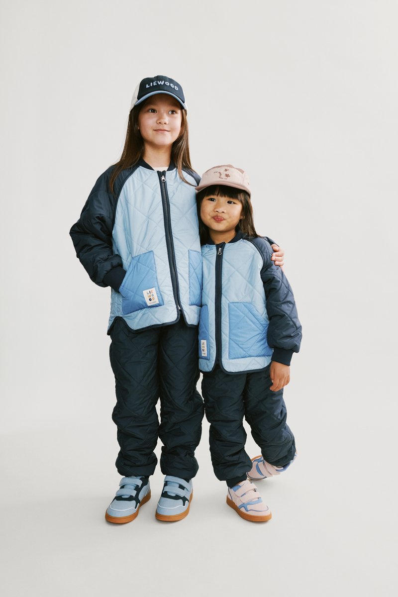 Δύο παιδιά φορούν συντονισμένα μπουφάν με καπιτονέ υφή σε ανοιχτό γαλάζιο και σκούρο ναυτικό μπλε, με αντίστοιχο σκούρο μπλε παντελόνι και πολύχρωμα παπούτσια. Καπέλα ολοκληρώνουν τις εμφανίσεις.