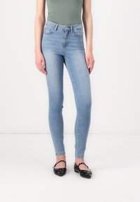 Woman wearing light blue skinny jeans, olive green fitted top, and black flats with strap and silver eyelets, standing against white background.