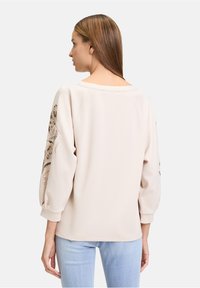 Sweatshirt beige clair avec des manches bouffantes 3/4, ornée d'accents floraux sur le bras gauche. Matière douce, coupe décontractée, encolure ras du cou.