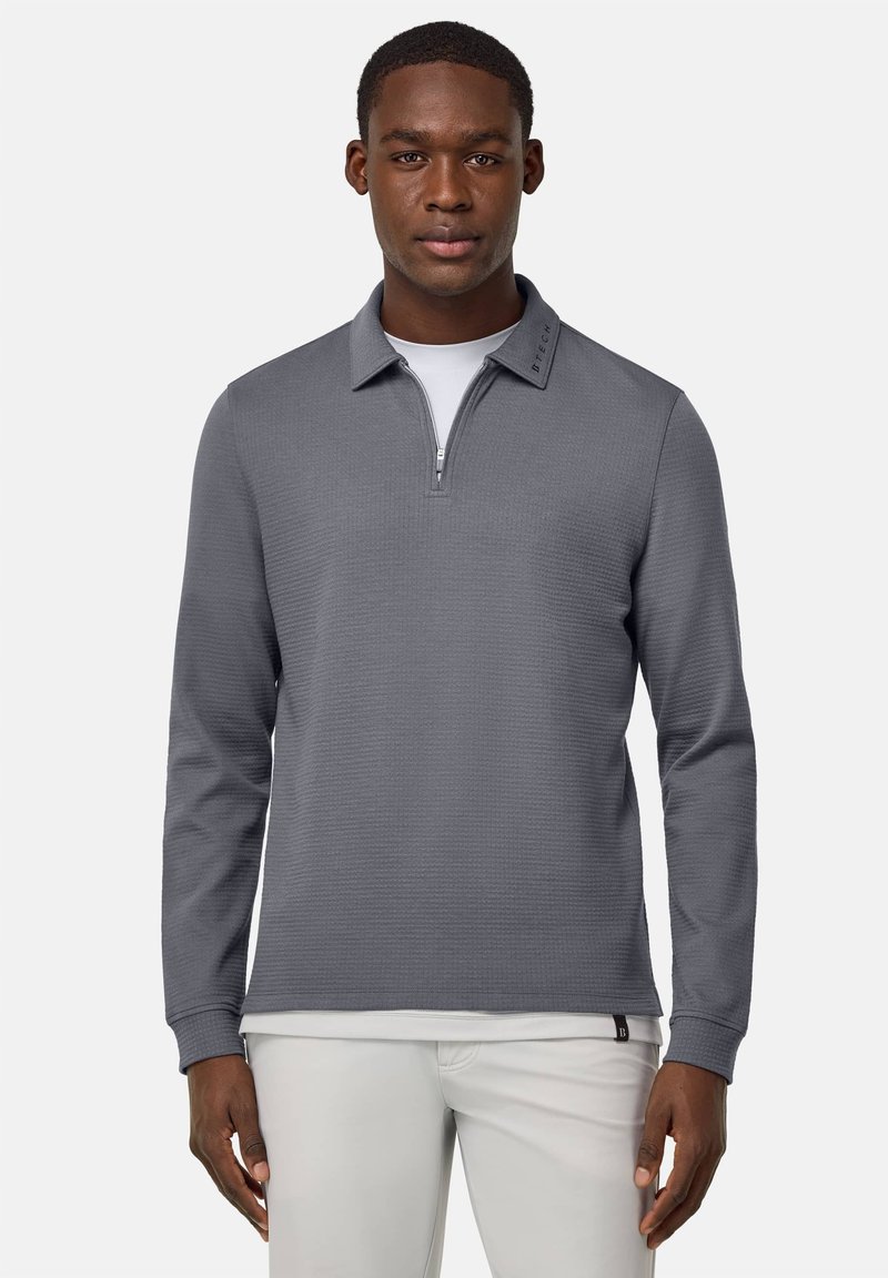 Pull-over gris à manches longues avec une fermeture éclair quart, en tissu texturé, et un col arborant un logo brodé. Porté par-dessus un T-shirt blanc, manches bien ajustées.