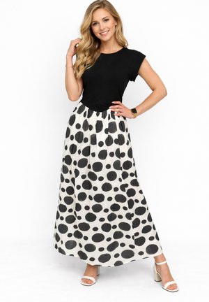 Mujer con cabello rubio largo que lleva una blusa negra de manga corta, falda larga blanca con grandes lunares negros, tacones blancos y un reloj, sonriendo.