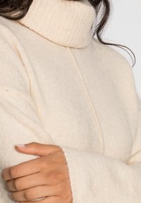 Beige turtleneck sweater with a ribbed collar, soft texture, dropped shoulders, and wide cuffs, showcasing a seam running down the front.