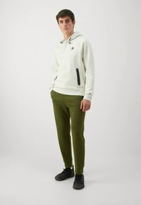 Sweatshirt branco com capuz e detalhes pretos, acompanhado de calças cónicas verde-oliva e sapatos desportivos pretos.