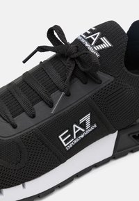 Zapatilla deportiva negra con parte superior de punto, cordones tonales y suela texturizada. Acentuada con un logotipo EA7 blanco en el lateral.
