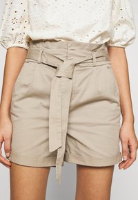 Beige high-waist shorts dengan sabuk kain lebar yang diikat di depan, dipasangkan dengan blus lengan puff putih dengan motif lubang.