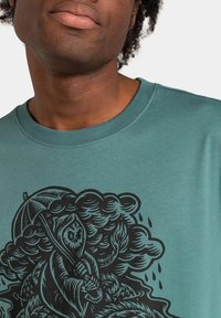 Camiseta de algodón verde azulado con un gran gráfico negro de una figura con capucha y un paraguas entre nubes y gotas de lluvia. Diseño de cuello redondo.