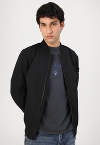 Black bomber jacket with a zip front, ribbed collar, and side pockets, layered over a gray t-shirt and paired with blue jeans.