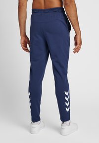 Marina joggers med en strukturerad yta, med dragsko i midjan och vita chevron-accenter på sidorna, i kombination med vita träningsskor.